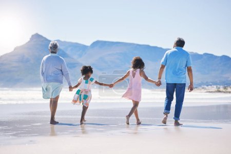 Photo for Back, holding hands and grandparents at the beach with children for holiday and walking by the sea. Content, summer and girl kids on a walk by the ocean with a senior man and woman for bonding. - Royalty Free Image