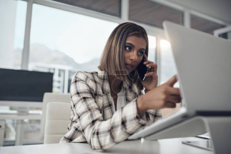 Photo for Im looking at the data right now. an attractive young businesswoman sitting alone in her office and using her cellphone while working on her laptop - Royalty Free Image