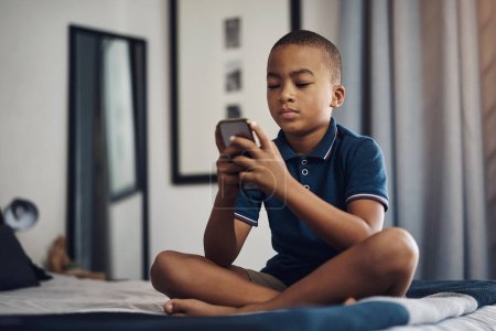 Foto de Utilizo mi tiempo de pantalla sabiamente. un niño usando un celular mientras está sentado en su cama - Imagen libre de derechos