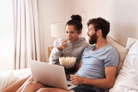 Téléchargez les photos : Transformer leur chambre en cinéma. un jeune couple heureux utilisant un ordinateur portable et mangeant du pop-corn tout en se relaxant sur le lit à la maison - en image libre de droit