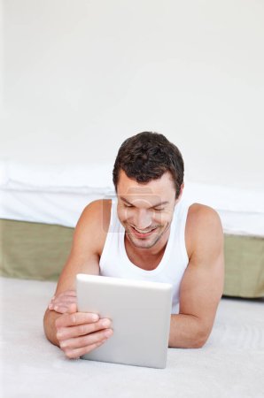Téléchargez les photos : Bénéficiant de la technologie d'écran tactile. Un bel homme allongé sur son lit travaillant sur sa tablette numérique - en image libre de droit