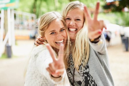 Foto de Sentir las vibraciones del festival. Retrato de dos jóvenes amigas rubias abrazando y dando la señal de paz mientras están afuera en un festival - Imagen libre de derechos
