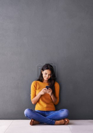 Foto de Uno de esos mensajes que te iluminan... una atractiva joven sentada en el suelo con su teléfono móvil - Imagen libre de derechos