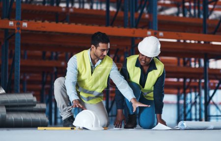Photo for Heres the plan, run with it. two young contractors crouching down in the warehouse together and using a blueprint to plan - Royalty Free Image