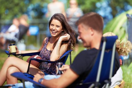 Photo for Portrait, woman drinking a beer at music festival or camping site and smiling with her friend. Vacation, happy female person sitting on camp chair and having a beverage while talking to her a male. - Royalty Free Image