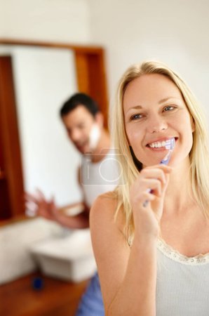 Photo for Enjoying each others company before going to work. A happy woman brushing her teeth while her boyfriend is in the background - Royalty Free Image