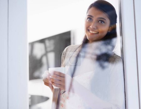 Foto de Disfrutando de una taza fresca. Una mujer joven mirando a través de un cristal mientras está de pie con una taza de café - Imagen libre de derechos