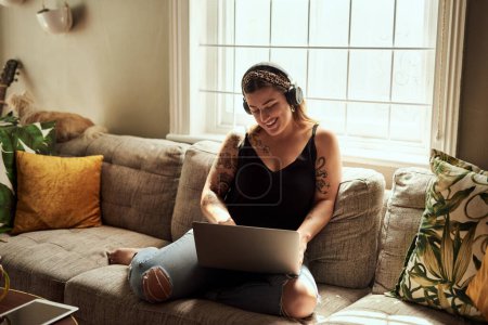 Photo for Computer, headphones and woman on sofa and music, happy work from home for mental health in apartment. Young person relax on couch and listening to audio technology, electronics and working on laptop. - Royalty Free Image
