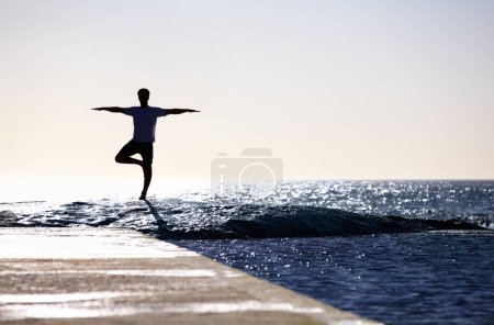 Téléchargez les photos : Homme, silhouette et yoga en méditation sur rocher de plage pour le bien-être spirituel, la paix intérieure ou le bien-être mental dans la nature. Yogi mâle dans l'arbre pose pour l'équilibre, le corps sain ou la pleine conscience sur l'océan. - en image libre de droit