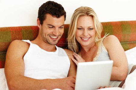 Foto de Esta tableta es increíble. Una atractiva pareja joven compartiendo una tableta digital mientras se relaja en la cama en casa - Imagen libre de derechos
