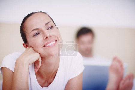 Téléchargez les photos : Une escapade romantique serait sympa... une jeune femme souriante allongée sur un lit pendant que son mari travaille sur un ordinateur portable en arrière-plan - en image libre de droit