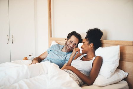 Téléchargez les photos : Couple interracial, rire et se détendre sur le lit pour les liens du matin, relation drôle ou blague à la maison. Femme heureuse et homme avec sourire, rire et couché dans la chambre à coucher pour parler ou converser à l'intérieur. - en image libre de droit
