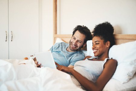 Téléchargez les photos : Couple heureux, tablette et se détendre sur le lit pour le divertissement du matin ou en streaming en ligne ensemble à la maison. Interracial homme et femme personne se détendre dans la chambre à coucher sur la technologie pour les médias sociaux ou la navigation. - en image libre de droit