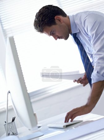 Foto de Comprobando la información en línea. Un joven hombre de negocios usando una computadora mientras revisa un documento - Imagen libre de derechos