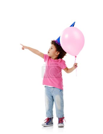 Foto de ¿Puedo jugar con eso? Estudio de una linda niña con un sombrero de fiesta y sosteniendo un globo sobre un fondo blanco - Imagen libre de derechos