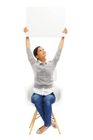 Foto de Esta idea va a ser enorme. Foto del estudio de una joven sentada en una silla con un cartel en blanco aislado en blanco - Imagen libre de derechos
