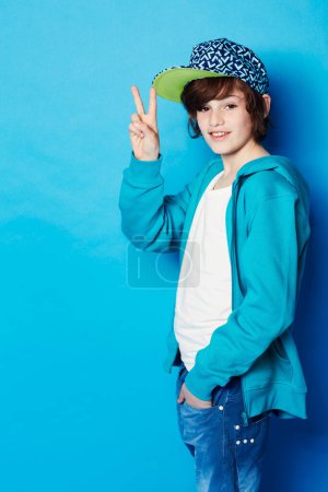 Photo for Fashion, peace sign and portrait of a boy in a studio with a casual, trendy and cool outfit. Teenager, smile and happy boy kid model with hipster apparel style posing isolated by a blue background - Royalty Free Image