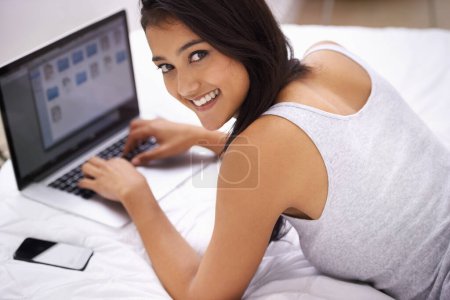 Téléchargez les photos : Elle n'est jamais loin de son ordinateur portable. Une jeune femme au lit avec son téléphone et son ordinateur portable - en image libre de droit