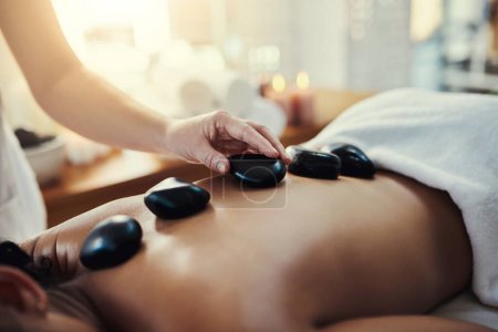Woman, hands and rocks for back massage at spa in beauty relaxation or skincare on bed. Hand of masseuse applying hot rock or stones on female for physical therapy, zen or skin treatment at resort.