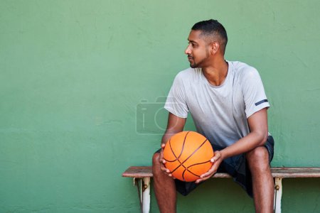 Foto de Me niego a perder. un joven deportivo sosteniendo una pelota de baloncesto mientras está sentado en un banco contra una pared - Imagen libre de derechos
