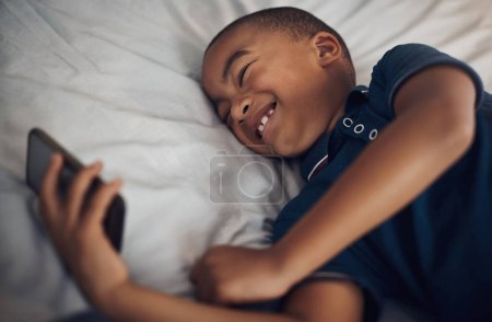 Foto de Eso es súper gracioso. un niño usando un celular mientras está acostado en su cama - Imagen libre de derechos