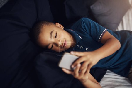 Foto de Depende de usted para controlar el uso de su teléfono inteligente de los niños. un niño usando un celular mientras está acostado en su cama - Imagen libre de derechos