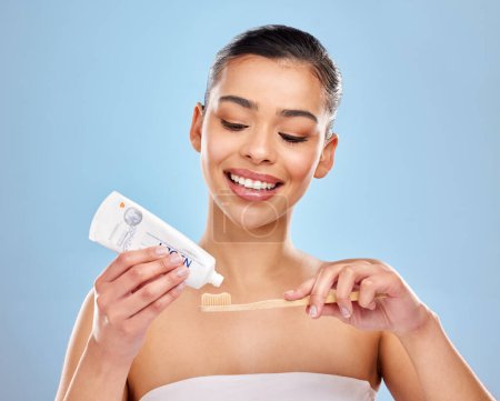 Téléchargez les photos : Utilisez un dentifrice qui contient du fluorure pour un sourire plus sain. Plan studio d'une jolie jeune femme appliquant du dentifrice sur une brosse à dents sur un fond bleu - en image libre de droit