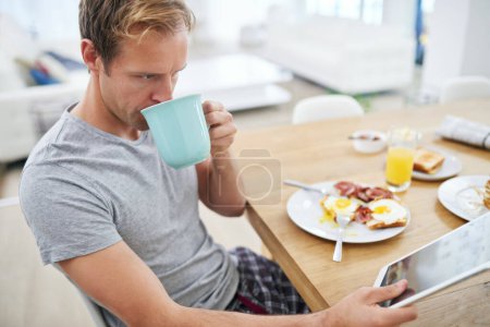 Foto de Tomando café y noticias. un hombre guapo usando su tableta mientras desayuna en casa - Imagen libre de derechos