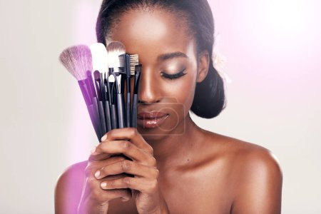 Téléchargez les photos : Beauté, outils de maquillage et femme noire avec brosses sur le visage en studio avec application cosmétique lueur. Soins de la peau, brosse et cosmétiques, modèle de soins du visage avec outil de contour de luxe sur fond blanc - en image libre de droit