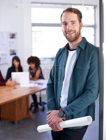 Photo for Hes a sought-after architect. Portrait of a young architect holding blueprints with his colleagues in the background - Royalty Free Image
