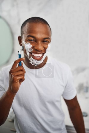 Téléchargez les photos : Homme noir, sourire et se raser avec lame, savon ou toilettage dans le miroir pour l'auto-soin dans la salle de bain à la maison. Africain mec, mousse ou crème pour l'épilation faciale, rasoir ou nettoyer pour l'hygiène le matin à la maison. - en image libre de droit