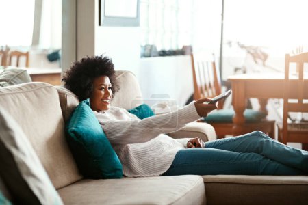 Photo for Black woman, watching tv and relax on couch with remote, home and happiness for me time, comedy or show. African girl, television and happy for movie, video or search on living room sofa in apartment. - Royalty Free Image