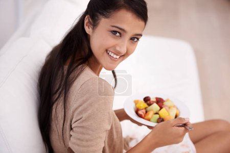 Téléchargez les photos : Fruits frais pour un mode de vie sain. Une belle jeune femme dégustant une collation saine dans le confort de son canapé - en image libre de droit