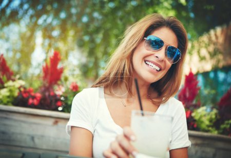 Foto de Qué bebida tan encantadora. una joven alegre con gafas de sol y disfrutando de una bebida fría mientras se sienta en un restaurante al aire libre durante el día - Imagen libre de derechos