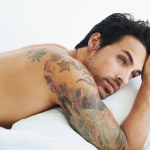 Thinking, relax and sexy with a topless man on a bed, lying in studio on a white background. Tattoo, idea and shirtless with a handsome young male model posing in a bedroom for sensuality or desire.