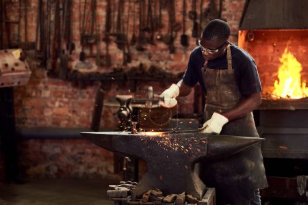 Forge forgeron, ouvrier et métal dans l'atelier et l'industrie manuelle travaillant sur acier chaud avec marteau, étincelles ou feu. Homme noir, fabrication d'outils de soudage ou de fer ou expert, commerce et espace de travail sombre.