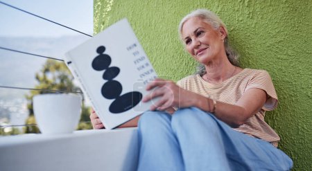 Téléchargez les photos : Les choses simples dans la vie. une séduisante femme âgée dégustant une tasse de café en lisant un livre à l'extérieur sur son balcon à la maison - en image libre de droit