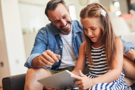 Téléchargez les photos : Il y a beaucoup à apprendre de papa. une adorable petite fille utilisant une tablette numérique avec son père sur le canapé à la maison - en image libre de droit