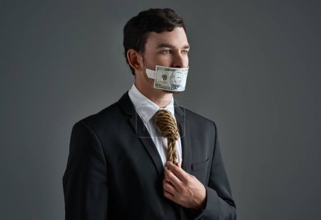 Foto de Where is the morality in the business market. Studio shot of a businessman with rope around his neck and money taped over his mouth against a gray background - Imagen libre de derechos