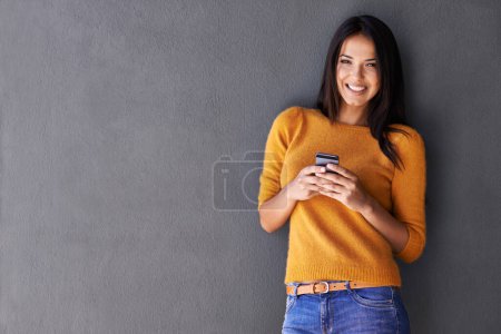 Téléchargez les photos : Rester en contact avec ses amis. Portrait d'une jolie jeune femme debout avec un téléphone portable contre un mur gris - en image libre de droit