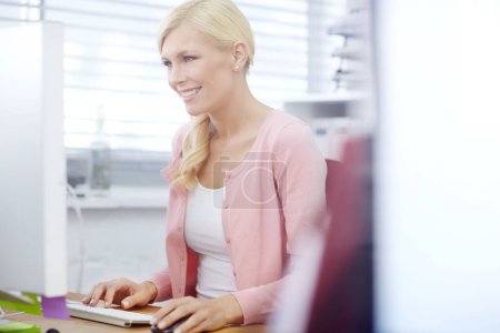 Téléchargez les photos : Une femme d'affaires utilisant un ordinateur de bureau à son bureau au travail. Une jeune travailleuse professionnelle tapant sur son clavier en regardant un écran et en souriant. Employé sympathique travaillant en ligne. - en image libre de droit