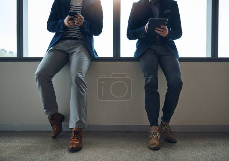 Foto de Ir en línea y hacer que el éxito suceda para usted. dos hombres de negocios que utilizan una tableta digital y un teléfono móvil mientras se apoyan contra una pared en una oficina moderna - Imagen libre de derechos