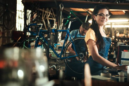 Téléchargez les photos : Portrait, sourire et femme travaillant dans un magasin de vélo pour réparation en magasin ou atelier de vélo. Visage, mécanicien de vélo et femme confiante, professionnelle ou technicienne avec bonheur et lunettes - en image libre de droit
