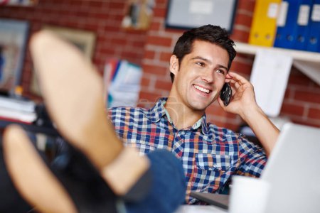 Foto de Hoy en día va a su manera. Un joven diseñador guapo recibiendo una llamada de un cliente mientras está sentado en su escritorio con los pies en alto - Imagen libre de derechos