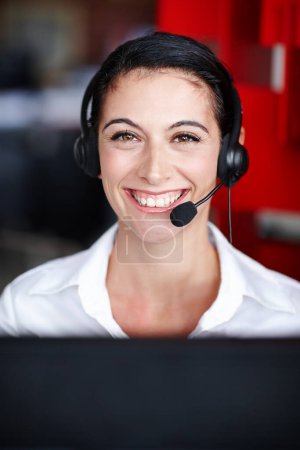Foto de Amistoso y diligente. Retrato de una bonita agente del centro de llamadas con auriculares y sentada frente a su computadora - Imagen libre de derechos