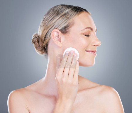 Téléchargez les photos : Les tonifiants peuvent aider à fermer les pores et à resserrer les trous cellulaires après le nettoyage. Plan studio d'une jolie femme mature utilisant un coton sur son visage sur un fond gris - en image libre de droit