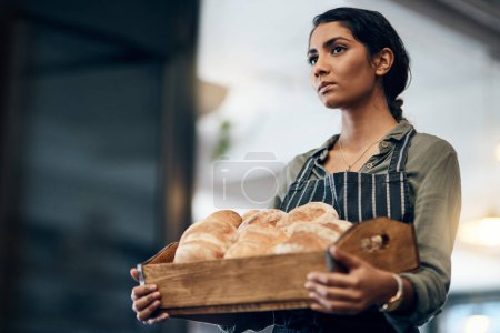 Téléchargez les photos : Servir quelques points de glucides sérieux. une jeune femme tenant une sélection de pains fraîchement cuits dans sa boulangerie - en image libre de droit