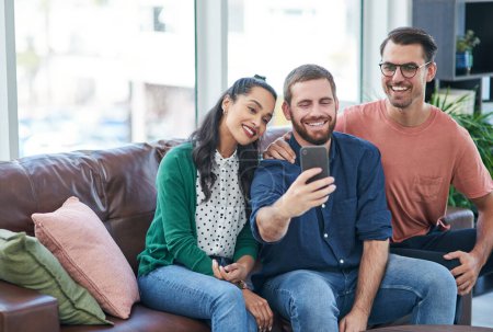 Téléchargez les photos : Les avantages de lancer une start-up avec des amis. deux jeunes hommes et une femme prenant des selfies ensemble sur un canapé - en image libre de droit