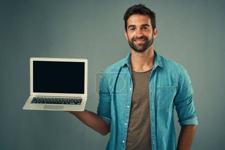 Foto de ¿Has visto el último zumbido en línea. Retrato de estudio de un joven guapo sosteniendo un portátil con una pantalla en blanco sobre un fondo gris - Imagen libre de derechos