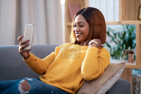 Téléchargez les photos : Femme noire dans sa maison, selfie et se détendre sur le canapé dans le salon, influenceur de style de vie et post sur les médias sociaux. Créatrice de contenu africaine dans l'appartement, sourire dans l'image et la communication avec l'application. - en image libre de droit